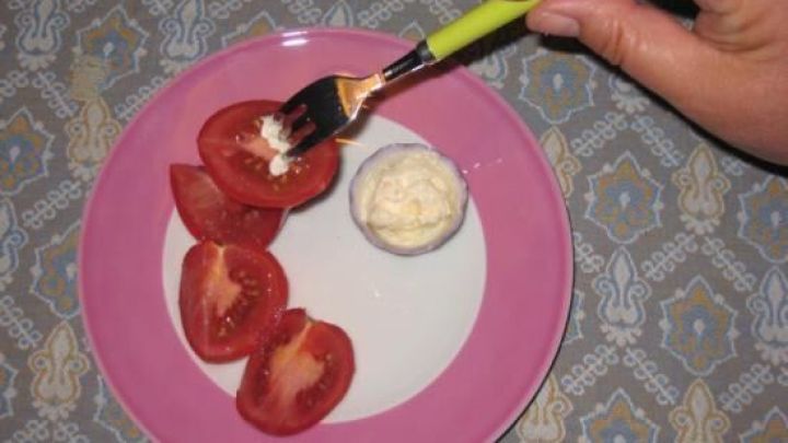 Hand h&auml;lt Gabel mit Mayonnaise &uuml;ber einer Tomatenscheibe auf einem Teller, platziert auf gemustertem Tisch.