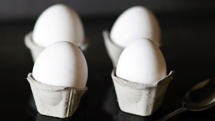 Selbstgemachte Einweg-Eierbecher aus Eierkartons halten wei&szlig;e Eier und stehen auf einer dunklen Oberfl&auml;che neben einem L&ouml;ffel.