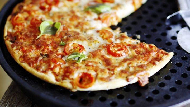 Eine frisch gebackene Pizza liegt auf einem schwarzen Gitterblech, zum Teil mit Kirschtomaten und Basilikum belegt.