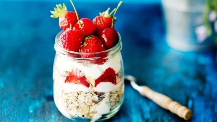 Ein Schraubglas enth&auml;lt ein M&uuml;sligemisch mit Joghurt und frischen Erdbeeren, auf einem leuchtend blauen Tisch, daneben steht eine Blumenvase.