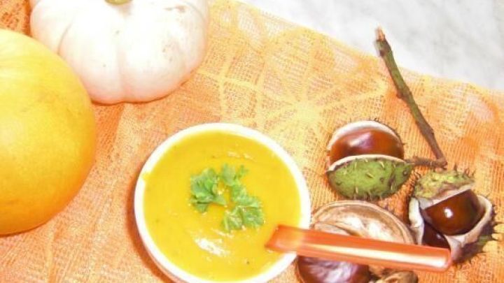 Eine Schale K&uuml;rbissuppe mit Petersilie, umgeben von Kastanien und K&uuml;rbissen auf einem orangen Tuch auf Marmor.