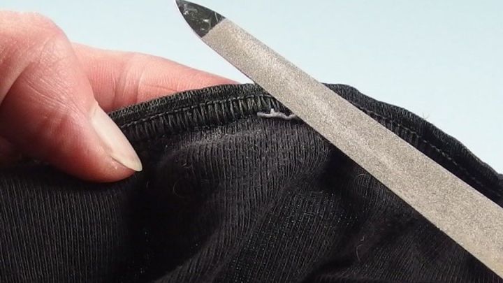 Eine Hand h&auml;lt ein Kleidungsst&uuml;ck, w&auml;hrend ein Werkzeug Piksende N&auml;hte entfernt, im Kontext eines hellen Hintergrunds.