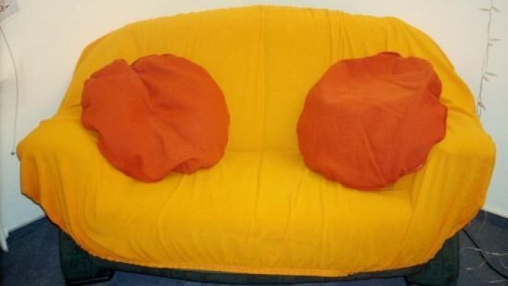 Ein altes Sofa ist mit einem leuchtend gelben Spannbettbezug bezogen und hat zwei orangefarbene Kissen darauf.