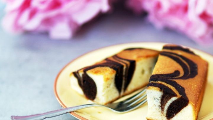 Ein saftiger Marmorkuchen mit Nougat und Amaretto liegt auf einem Teller, umgeben von zarten, rosa Bl&uuml;ten im Hintergrund.
