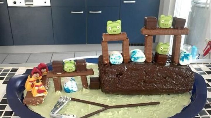 Ein bunter Angry Birds Kuchen steht auf einem Teller, umgeben von Spielzeugfiguren und K&uuml;chenutensilien in einer modernen K&uuml;che.