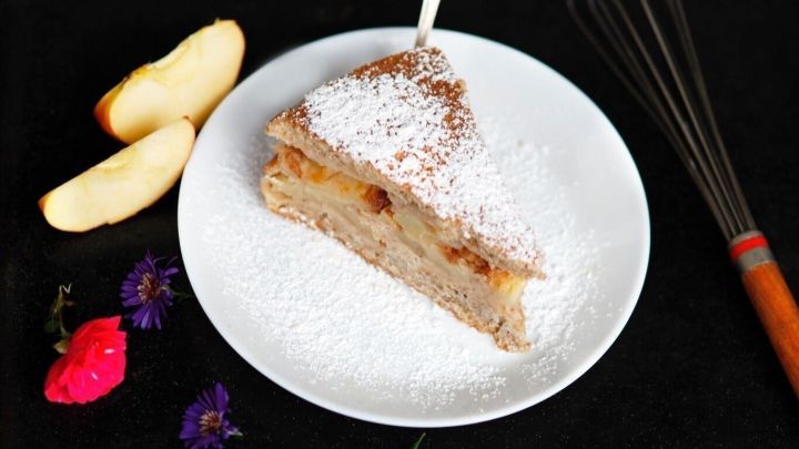 Ein St&uuml;ck versunkener Apfelkuchen liegt auf einem Teller, umgeben von &Auml;pfeln und bunten Blumen auf schwarzem Hintergrund.