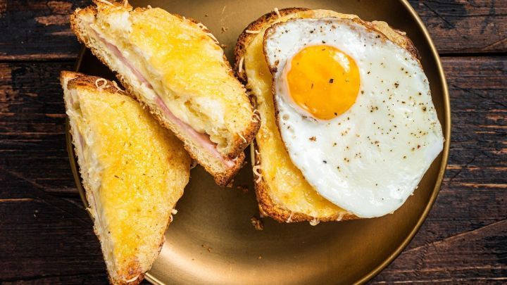 Ein golden gebratens Fr&uuml;hst&uuml;cksei liegt auf ger&ouml;stetem Brot, umgeben von geschmolzenem K&auml;se und Schinken.