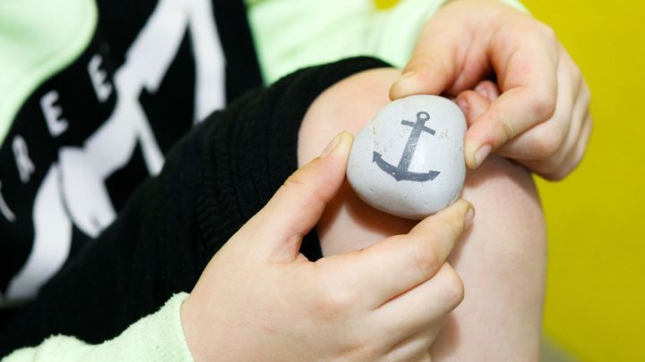 Ein Kind h&auml;lt einen k&uuml;hlen Stein mit einem Anker-Motiv gegen das Knie, um ihn als Coolpack zu nutzen.