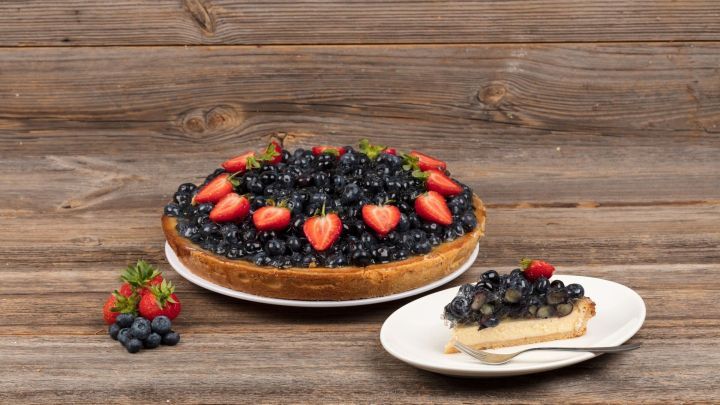 Ein Blaubeerk&auml;sekuchen mit frischen Erdbeeren, angerichtet auf einem Holzuntergrund, wartet auf eine k&ouml;stliche Verkostung.