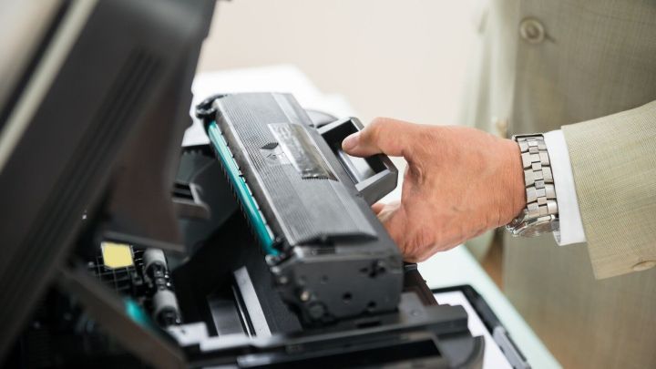 Ein Hand greift eine Tonerkartusche und setzt sie in einen Drucker ein, w&auml;hrend die Umgebung hell und modern wirkt.