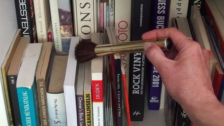 Ein Make-up-Pinsel wird verwendet, um Staub von B&uuml;chern in einem Regal zu wischen.