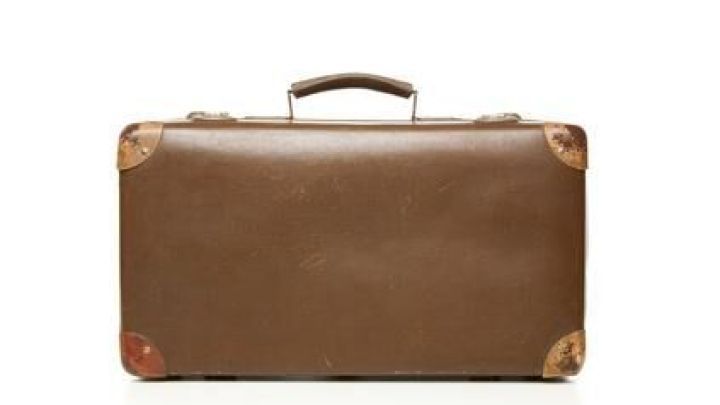 Ein alter brauner Koffer steht geschlossen vor einem wei&szlig;en Hintergrund, zeigt abgenutzte Ecken und einen Griff oben.