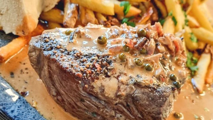 Zartes Steak liegt in cremiger Sauce auf einem Teller, umgeben von knusprigen Pommes und Brot.