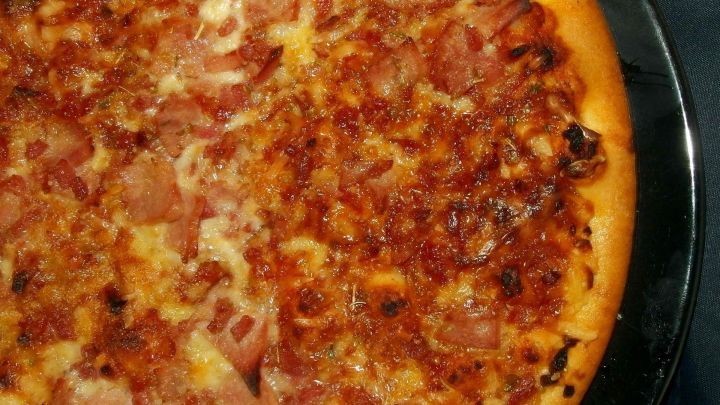 Eine frisch gebackene Pizza liegt auf einem schwarzen Teller, der Belag besteht aus K&auml;se und Schinken, appetitlich und knusprig.