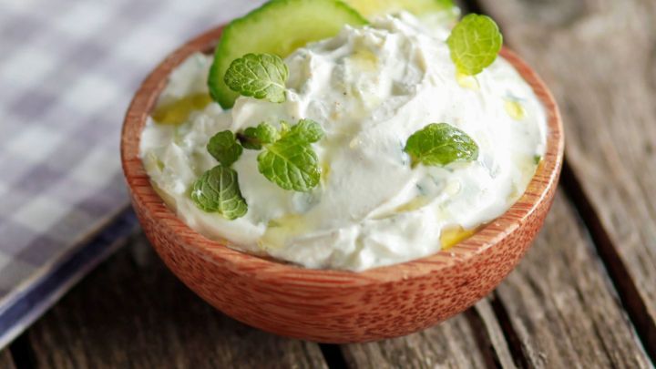 Eine Holzschale mit frischem Cacik, garniert mit Gurkenscheiben und Minzbl&auml;ttern, steht auf einem rustikalen Holztisch.