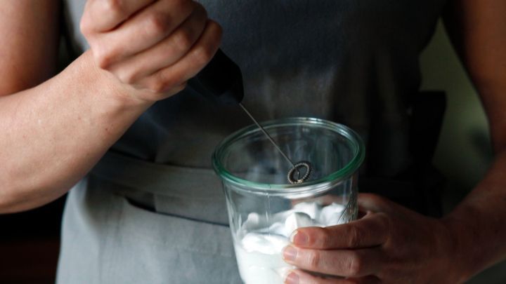 Eine Person schl&auml;gt Eiwei&szlig; mit einem Milchaufsch&auml;umer in einem transparenten Gef&auml;&szlig; in einer k&uuml;chen&auml;hnlichen Umgebung.