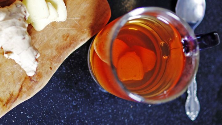 Eine Tasse Ingwer-Salbei-Honig-Tee steht auf einem dunklen Tisch, daneben liegt frischer Ingwer, umgeben von einer L&ouml;ffel.