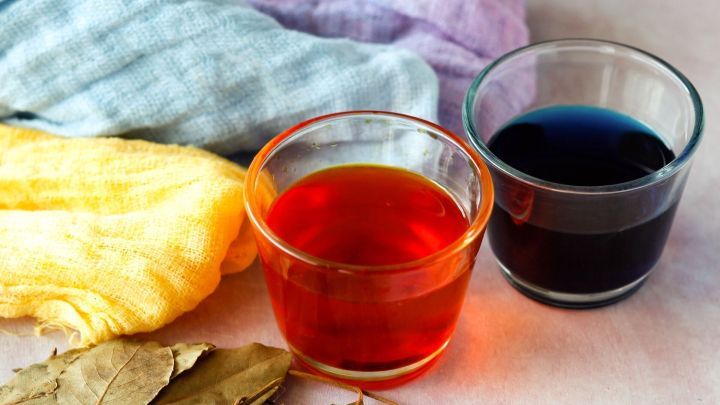 Bunte Fl&uuml;ssigkeiten in Gl&auml;sern stehen neben farbigen Stoffen und getrockneten Bl&auml;ttern, ideal zum F&auml;rben mit Pflanzenfarbe.