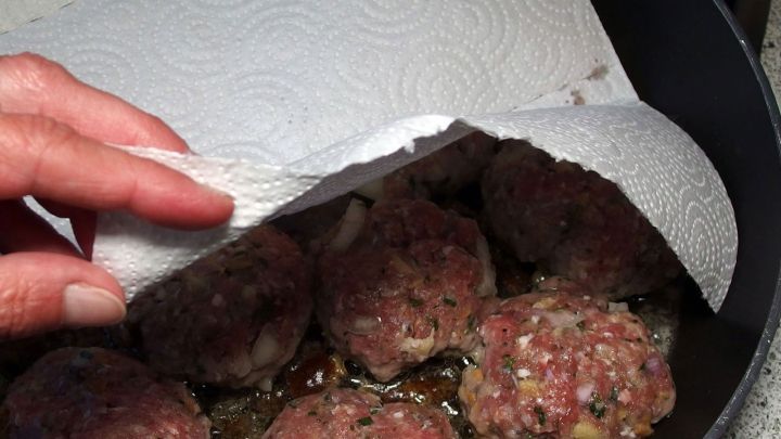 Der K&uuml;chenkrepp wird von einer Hand angehoben, w&auml;hrend fleischige B&auml;llchen in einer Pfanne braten, um den Herd sauber zu halten.