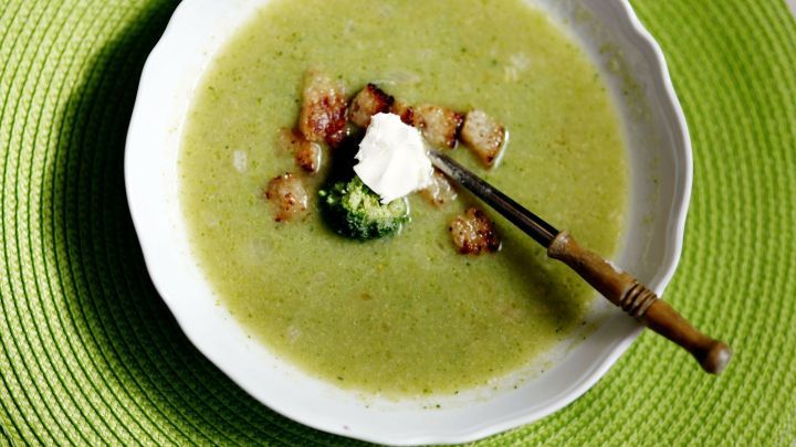 Suppe mit Croutons und einem Klecks Sahne in wei&szlig;er Sch&uuml;ssel, auf gr&uuml;nem Platzdeckchen, neben einem Holzl&ouml;ffel.