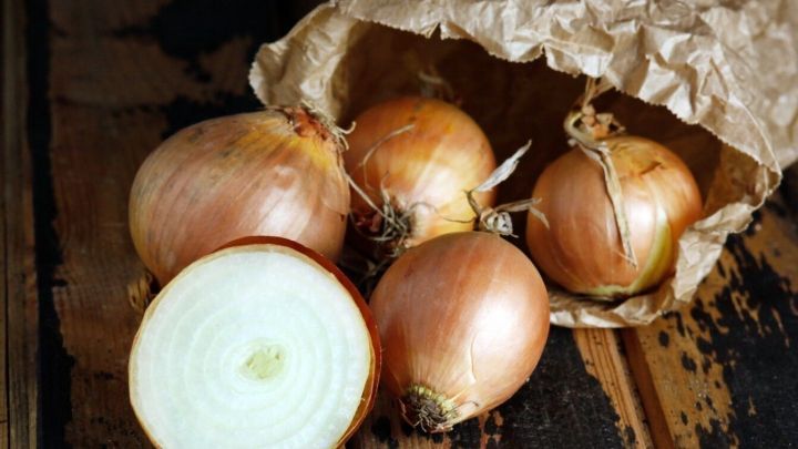 Eine braune Papiert&uuml;te liegt offen und zeigt mehrere Zwiebeln, w&auml;hrend eine halbierte Zwiebel frischen Zwiebelgeruch verbreitet.