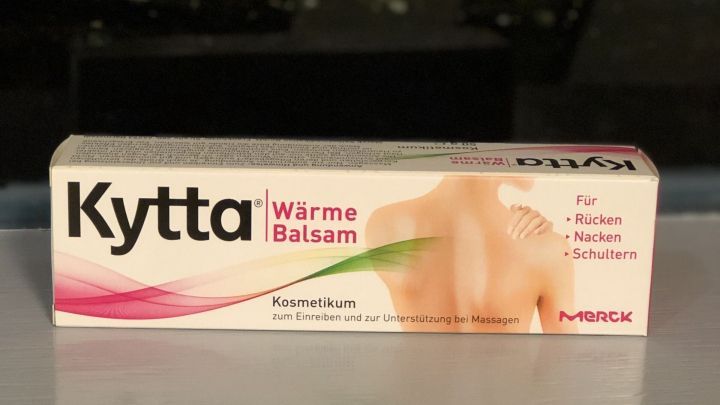 Eine Verpackung von Kytta W&auml;rmbalsam steht auf einem Tisch, w&auml;hrend im Hintergrund gr&uuml;ne Pflanzen sichtbar sind.