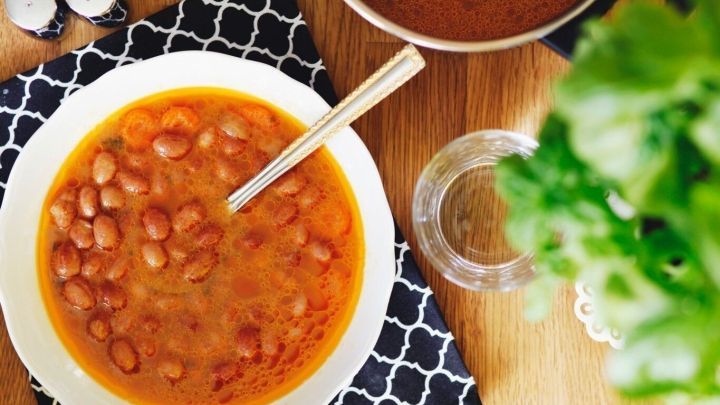 Eine Sch&uuml;ssel Ostfriesische Bohnensuppe mit Bohnen und Gem&uuml;sebr&uuml;he steht auf einem Tisch, umgeben von Gew&uuml;rzen und einer Pflanze.