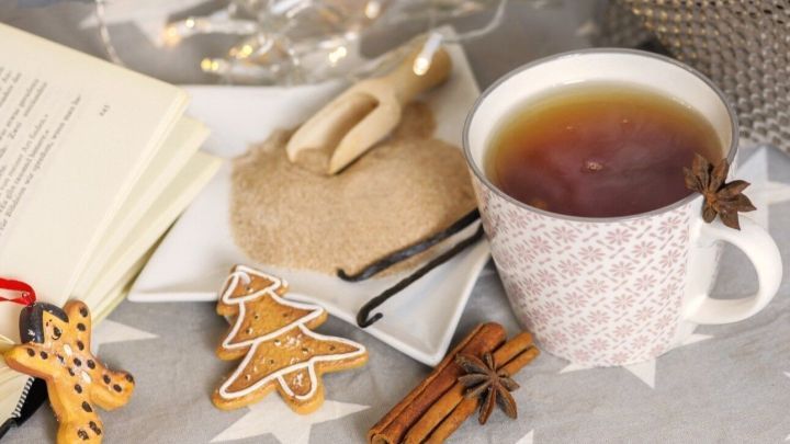 Eine Tasse Gew&uuml;rztee steht auf einem dekorativen Tisch neben Keksen, Zucker und einer offenen Buchseite im Hintergrund.