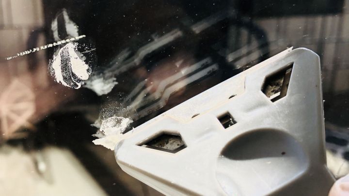 Ein Glaskeramikschaber entfernt Farbspritzer von einem Fensterglas in einem hellen Innenraum mit reflektierenden Oberfl&auml;chen.