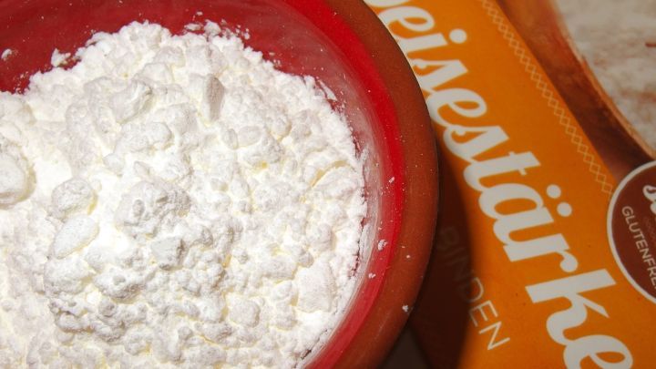 Ein roter Sch&uuml;ssel mit St&auml;rkemehl steht auf einer Holzoberfl&auml;che neben einer Verpackung. Der Mehl wirkt fein und frisch.