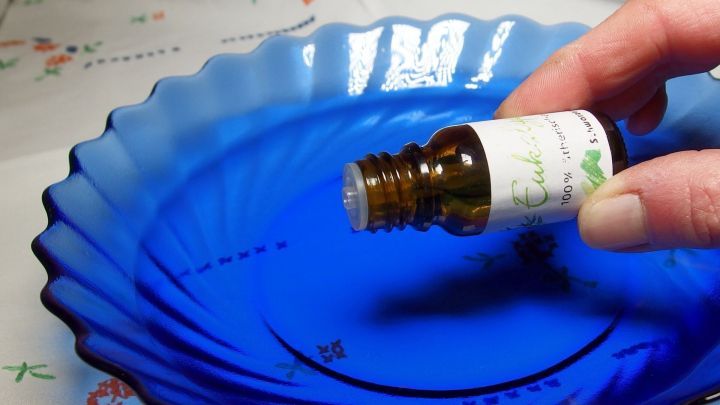 Eine Hand h&auml;lt eine Flasche Eukalyptus&ouml;l &uuml;ber einer blauen Schale, bereit, es in hei&szlig;es Wasser zu geben.