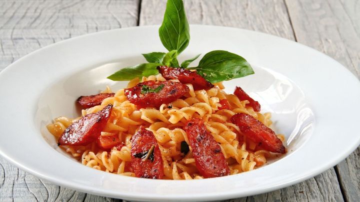 Ein Teller mit Nudeln wird mit knuspriger Mettwurst und frischem Basilikum angerichtet, auf einem rustikalen Holztisch.