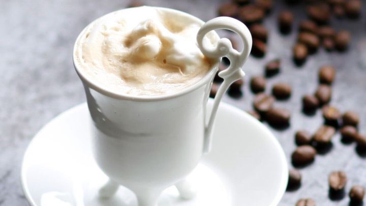Eine stilvolle Tasse mit cremigem Kaffee steht auf einem Teller, umgeben von Kaffeebohnen auf einer eleganten Oberfl&auml;che.