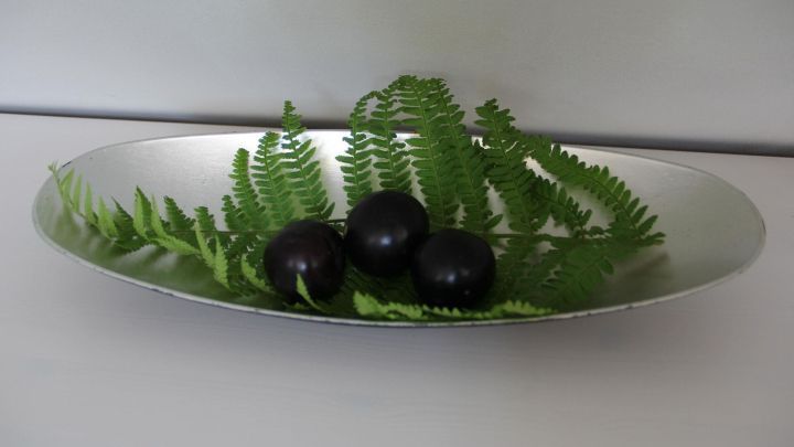 Drei schwarze Fr&uuml;chte liegen auf einem gr&uuml;nen Farnblatt in einer silbernen Schale, harmonisch angerichtet.