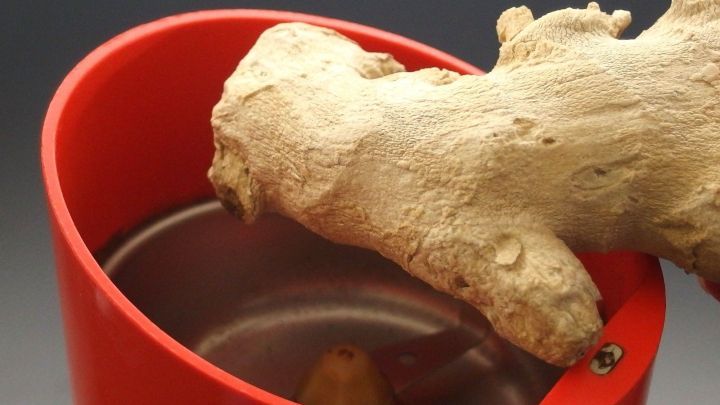 Ein St&uuml;ck frischer Ingwer wird &uuml;ber einer elektrischen Kaffeem&uuml;hle gehalten, die in einem roten Beh&auml;lter untergebracht ist.