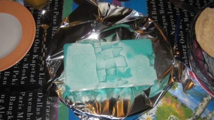 Eine K&auml;seplatte, eingewickelt in Folie, wird auf einem bunten Tisch mit anderen Speisen serviert.