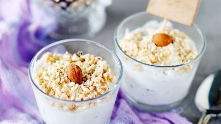 Zwei Dessertgl&auml;ser mit crunchy Toppings stehen auf einem lila Tuch, umgeben von N&uuml;ssen und einem dekorativen Sch&auml;lchen.