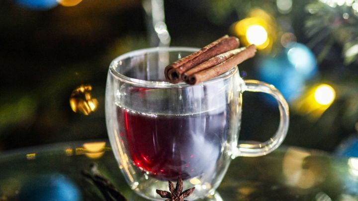 Ein Glas Gl&uuml;hwein mit Zimtstangen steht auf einem Tisch, umgeben von Weihnachtslichtern und dekorationen.