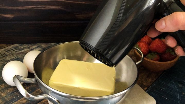 Ein F&ouml;hn erw&auml;rmt ein St&uuml;ck kaltes Backfett in einer Sch&uuml;ssel, um es f&uuml;r das Backen vorzubereiten. Im Hintergrund sind Eier und Fr&uuml;chte sichtbar.