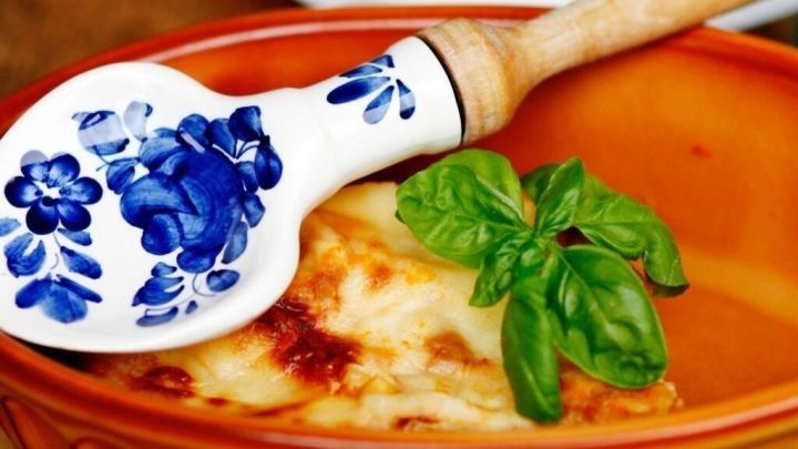 Eine goldbraun gebackene Lasagne liegt in einer terrakottafarbenen Sch&uuml;ssel, umgeben von einem L&ouml;ffel mit blauer Verzierung und frischem Basilikum.
