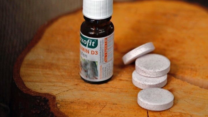 Eine Flasche mit Vitamin D und mehrere Tabletten liegen auf einem Holzst&uuml;ck, umgeben von einem ruhigen Naturhintergrund.