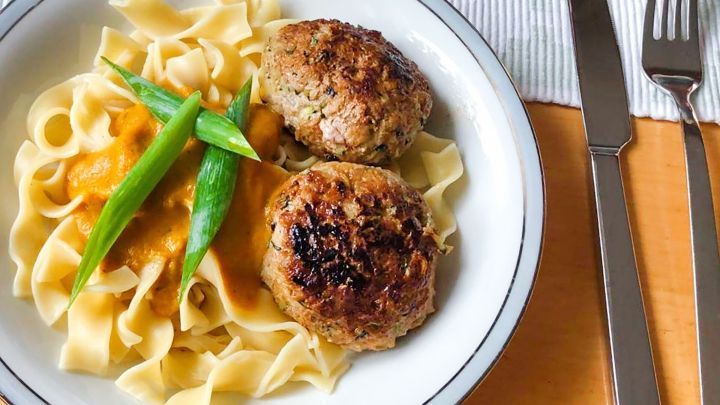 Zwei knusprige Frikadellen liegen auf einem Teller mit Bandnudeln und frischen Erbsen, serviert auf einem gedeckten Tisch.