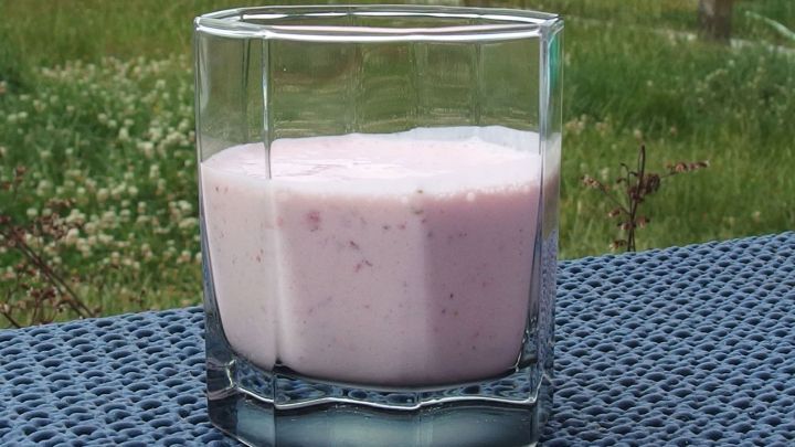 Ein Glas Erdbeer-Quark steht auf einem Tisch im Freien und zeigt eine cremige, rosa Masse vor einer gr&uuml;nen Wiese.