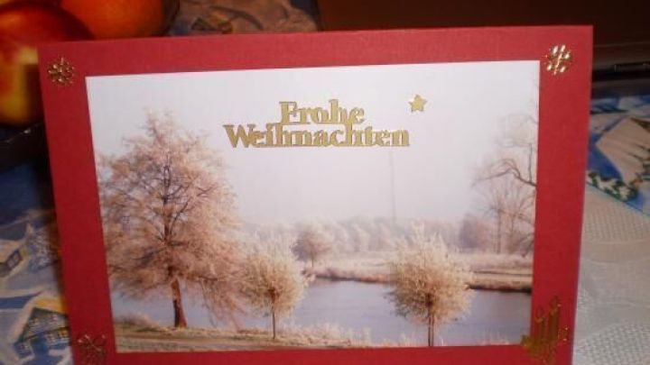Eine rote Weihnachtskarte steht auf einem Tisch und zeigt eine winterliche Landschaft mit B&auml;umen im Schnee. Text: "Frohe Weihnachten".