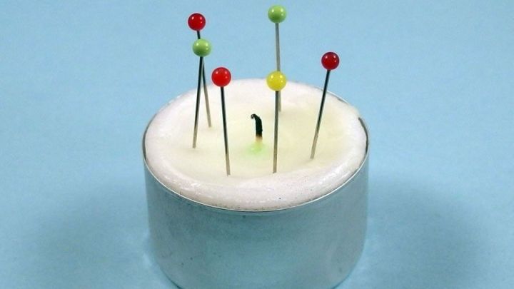 Ein Teelicht dient als improvisierter Nadelhalter, mit bunten Nadeln, die sicher darin stecken, auf einem hellblauen Hintergrund.