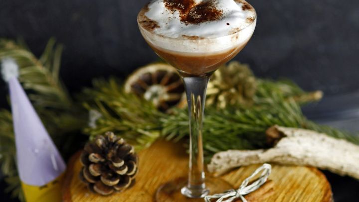 Ein Glas Weihnachtslik&ouml;r steht auf einem rustikalen Holzbrett, umgeben von Weihnachtsdekoration und Tannenzweigen.