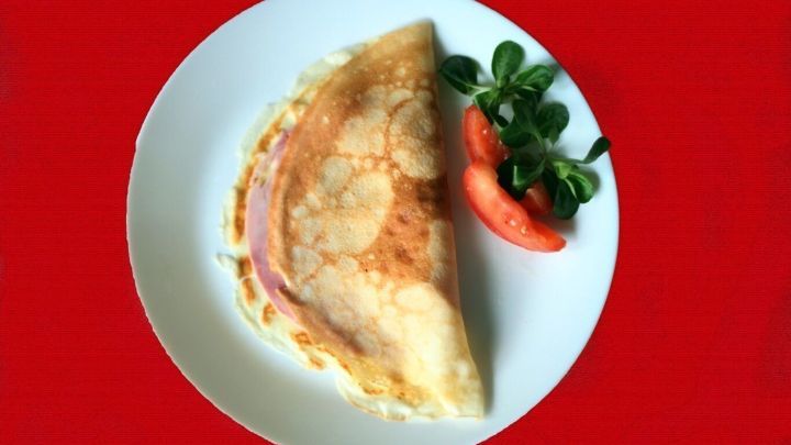 Auf einem wei&szlig;en Teller liegt eine in zwei H&auml;lften gefaltete Pfannkuchen-Calzone, umgeben von einer Tomatenscheibe und frischem Salat.