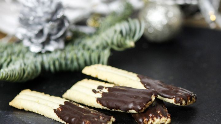 Die Spritzgeb&auml;ck-Variante liegt auf einem schwarzen Teller, teilweise mit Schokolade &uuml;berzogen, umgeben von festlichen Dekorationen.