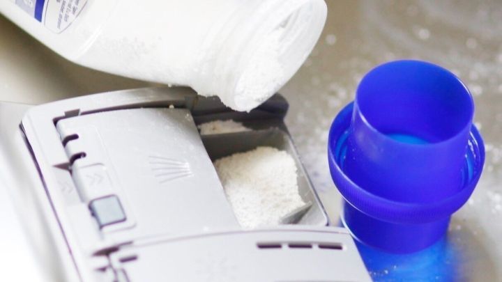 Eine Flasche Waschpulver sch&uuml;ttet Pulver in das Fach der offenen Sp&uuml;lmaschine, um diese zu s&auml;ubern.