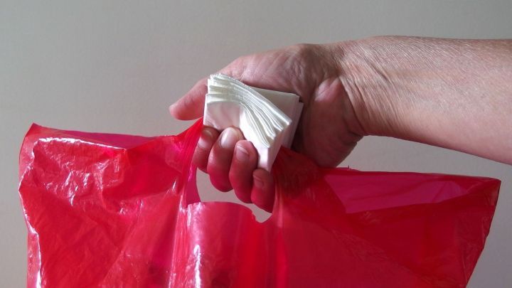 Eine Hand h&auml;lt eine rote Plastikt&uuml;te mit einem geknoteten Griff, der anzeigt, dass sie gef&uuml;llt und bereit zum Tragen ist.